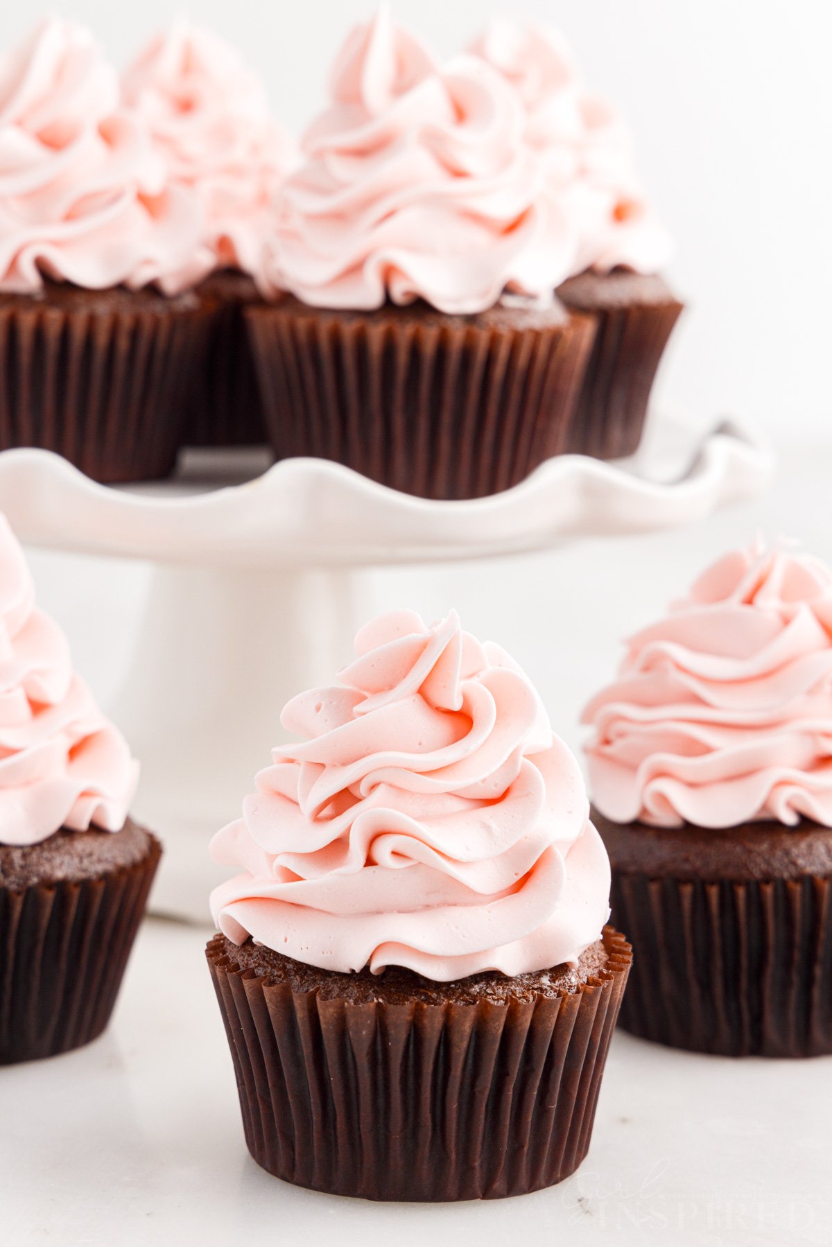 Chocolate cupcake with big swirl of pink strawberry frosting, with cake platter full of cupcakes in background.