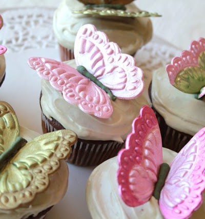 molded gumpaste butterflies in pink and green, on top of white frosting topped cupcakes