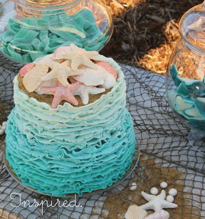 blue wavy ocean themed cake with candy seashells on top, sitting on fish net with jars with shark candy in background