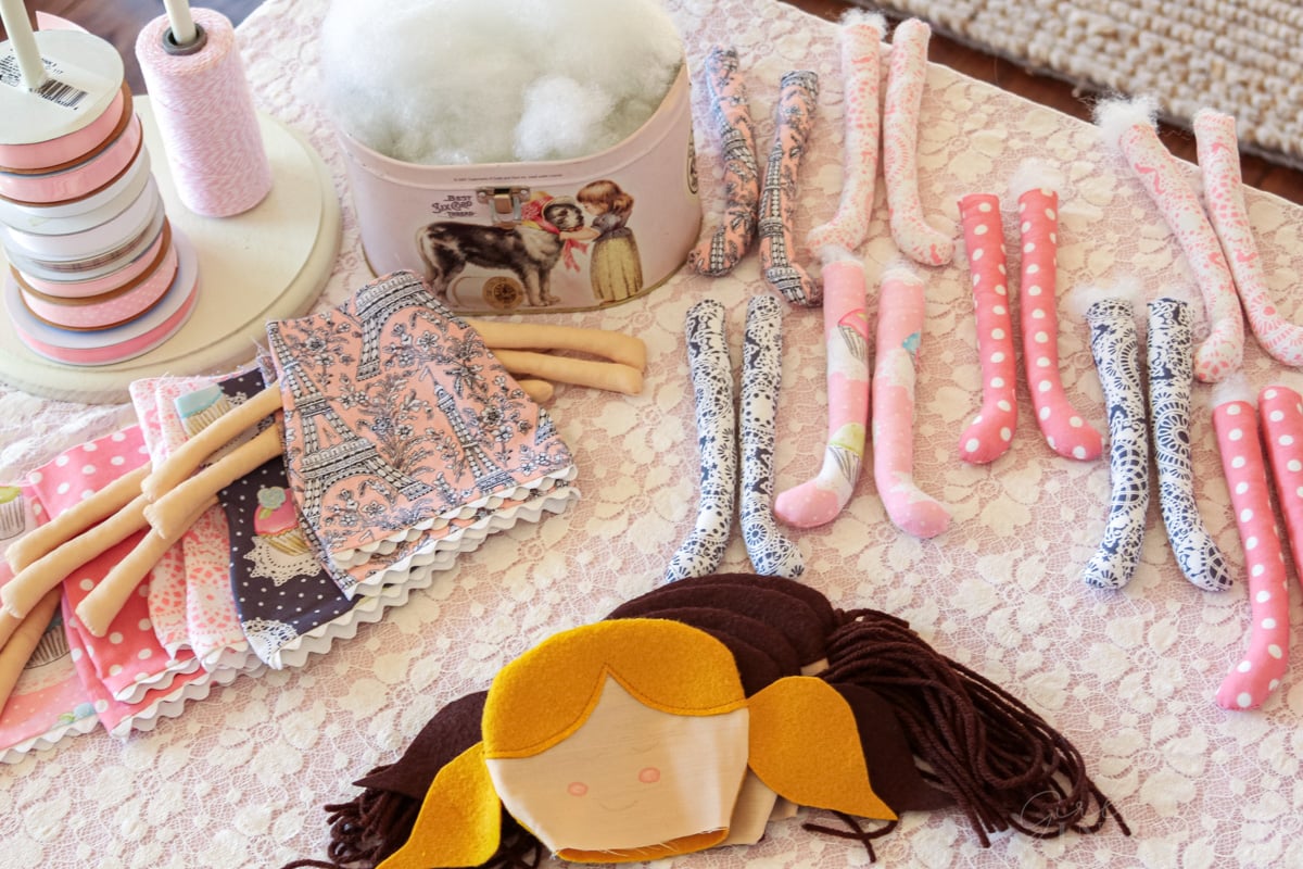 A variety of doll heads, doll body and arm pieces, and pairs of doll legs set on a table with fabric stuffing and spools of ribbon.