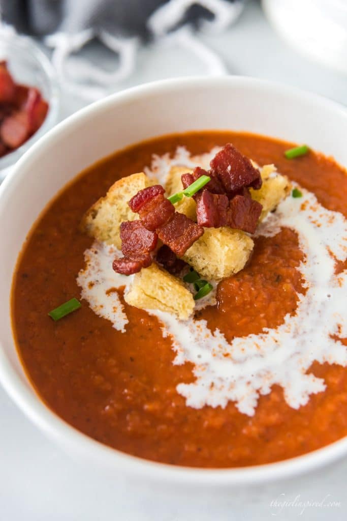 tomato bisque in white bowl with bacon, croutons, chives and drizzle of cream on top