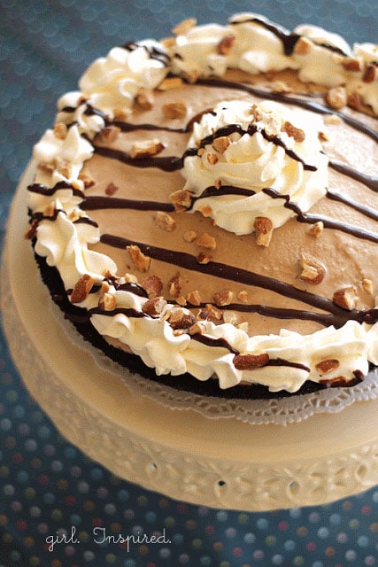 mud pie with whipped cream border and chocolate drizzle/toasted almonds on top, sitting on doily and cream cake stand on blue polka dot tablecloth