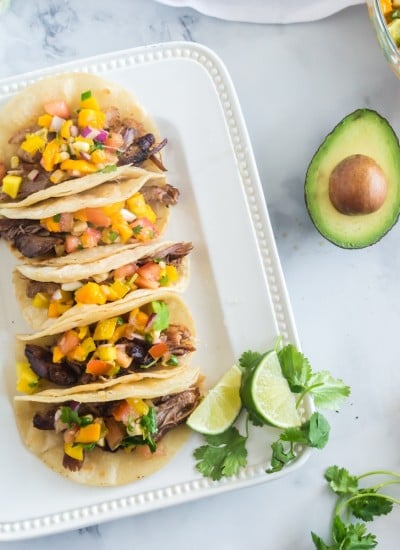 overhead view of crockpot carnitas tacos on white plate with mango salsa, lime wedge, avocado, and cilantro