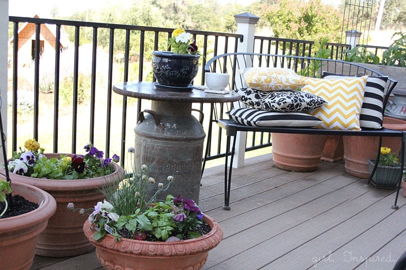 Wooden top outdoor side table with antique milk can base, blue pot of flowers and white mug on table, black bench with yellow and black throw pillows, potted flowers on deck