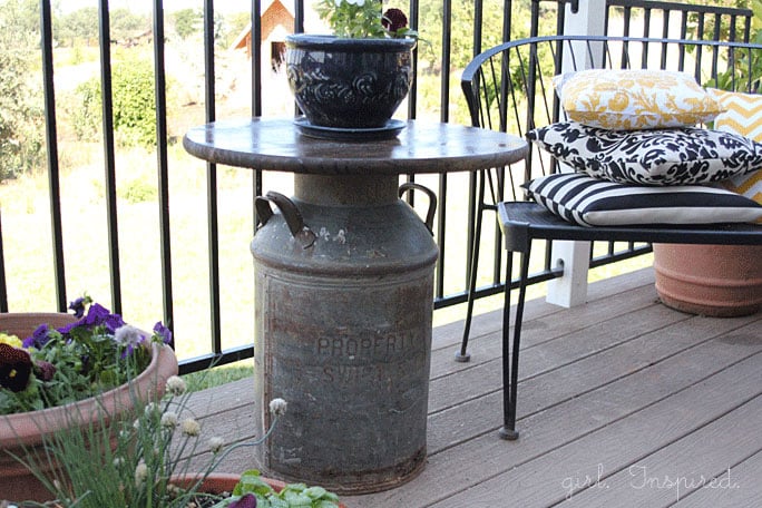 Wooden top outdoor side table with antique milk can base, blue pot of flowers and white mug on table, black bench with yellow and black throw pillows, potted flowers on deck
