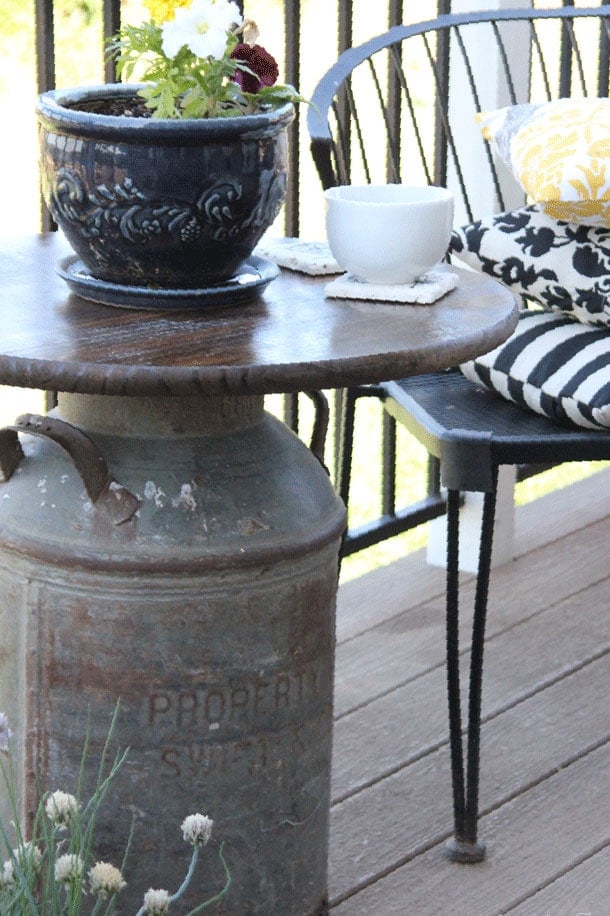 Wooden top outdoor side table with antique milk can base, blue pot of flowers and white mug on table, black bench with yellow and black throw pillows