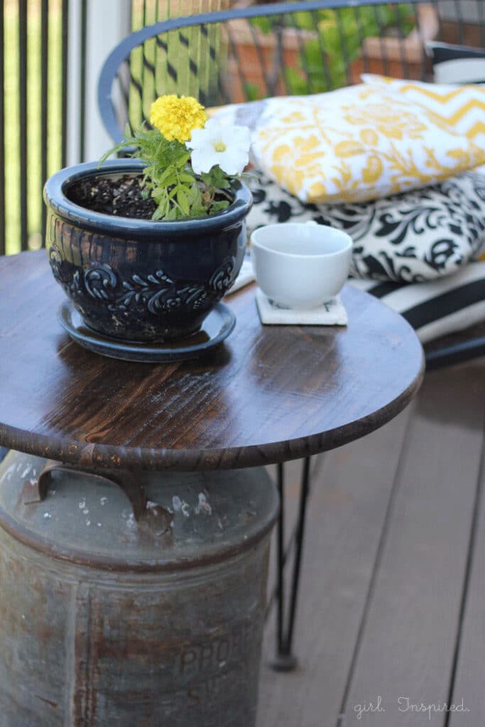Wooden top outdoor side table with antique milk can base, blue pot of flowers and white mug on table, black bench with yellow and black throw pillows