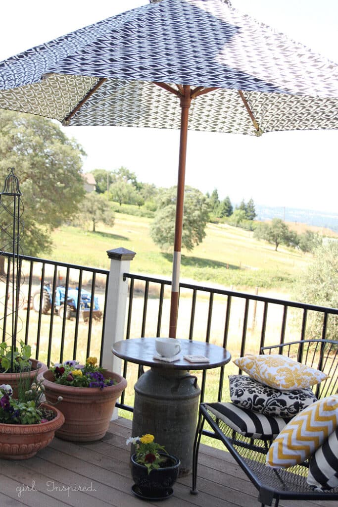 Wooden top outdoor side table with antique milk can base, blue pot of flowers and white mug on table, black bench with yellow and black throw pillows, potted flowers on deck, and black and white umbrella in milk can side table center