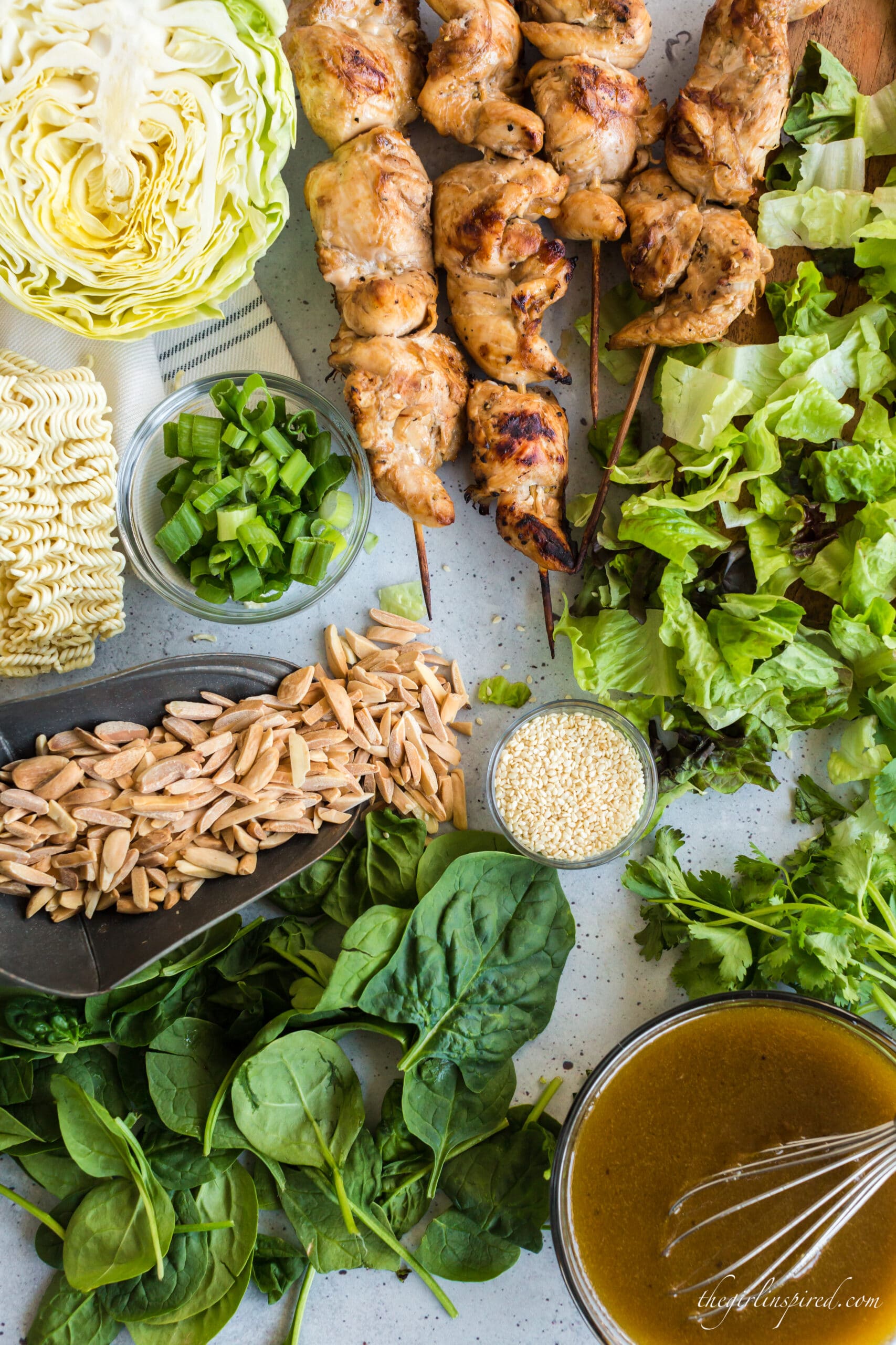 overhead view of chinese chicken salad ingredients: spinach, lettuce, green onions, almond slivers, top ramen noodles, cabbage, sesame seeds, cilantro, and bowl of dressing with whisk