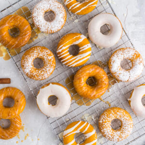 overhead of two ingredient pumpkin donuts on cookie sheet with cinnamon sticks and a variety of toppings - powdered sugar, white frosting, caramel drizzle