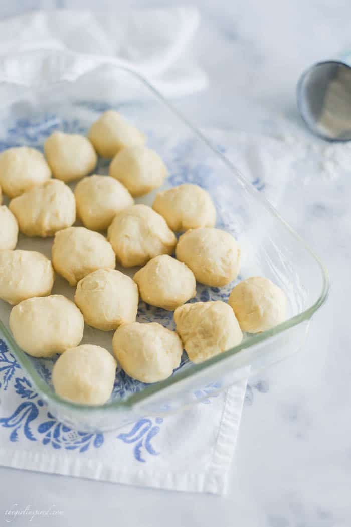overhead view of raw dinner roll dough balls in clear glass baking dish with blue floral linen below and blue cup with flour