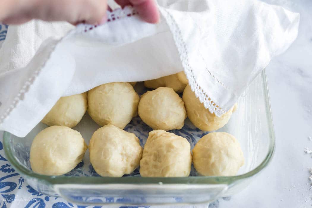 raw dinner roll dough balls in clear glass baking dish with blue floral linen below and white dish towel over the top