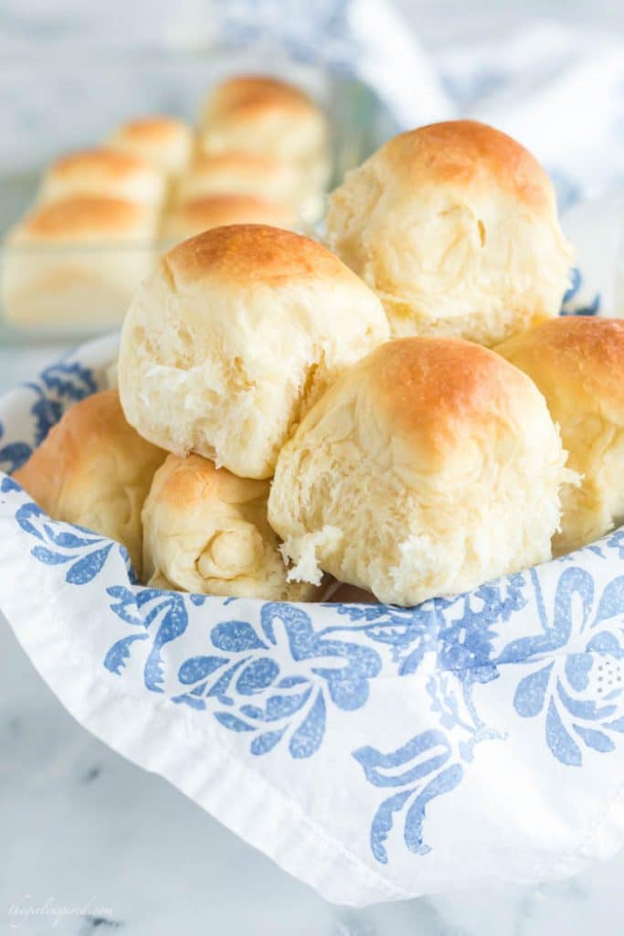 dinner rolls stacked in a bread basket with white and blue floral linen, baking dish with rolls