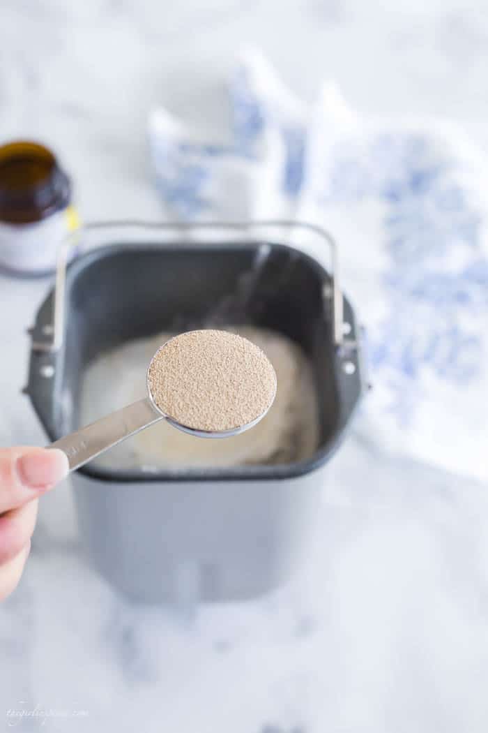 spoonful of yeast in silver measuring spoon above bread machine pan filled with ingredients