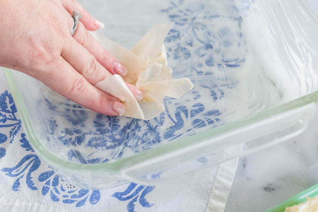 hand using parchment paper to spread butter in clear baking dish with blue floral dish towel