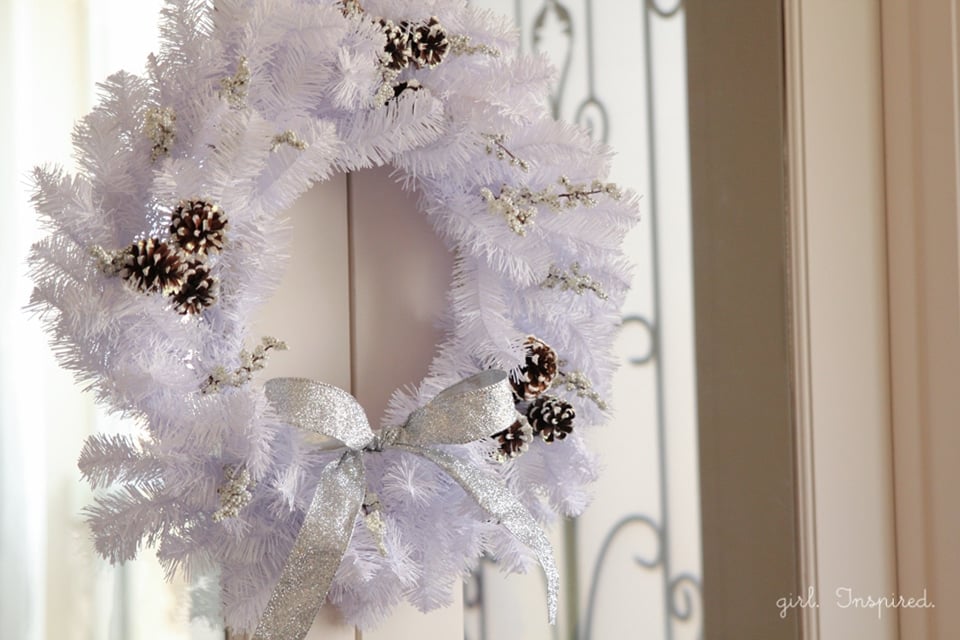 A white wreath dusted with snow, white berry sprigs, snow tipped pinecones and a silver glitter ribbon bow on a glass door