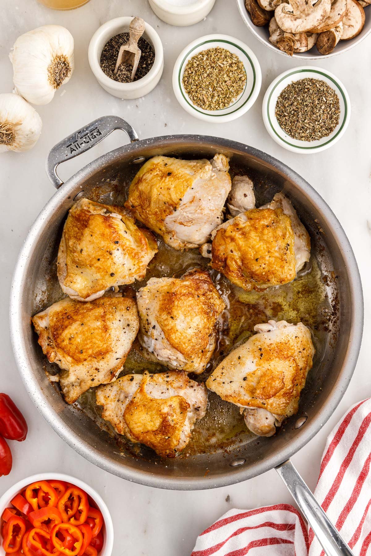 Browned chicken thighs in large skillet with red striped linen and other ingredients prepped in small bowls.