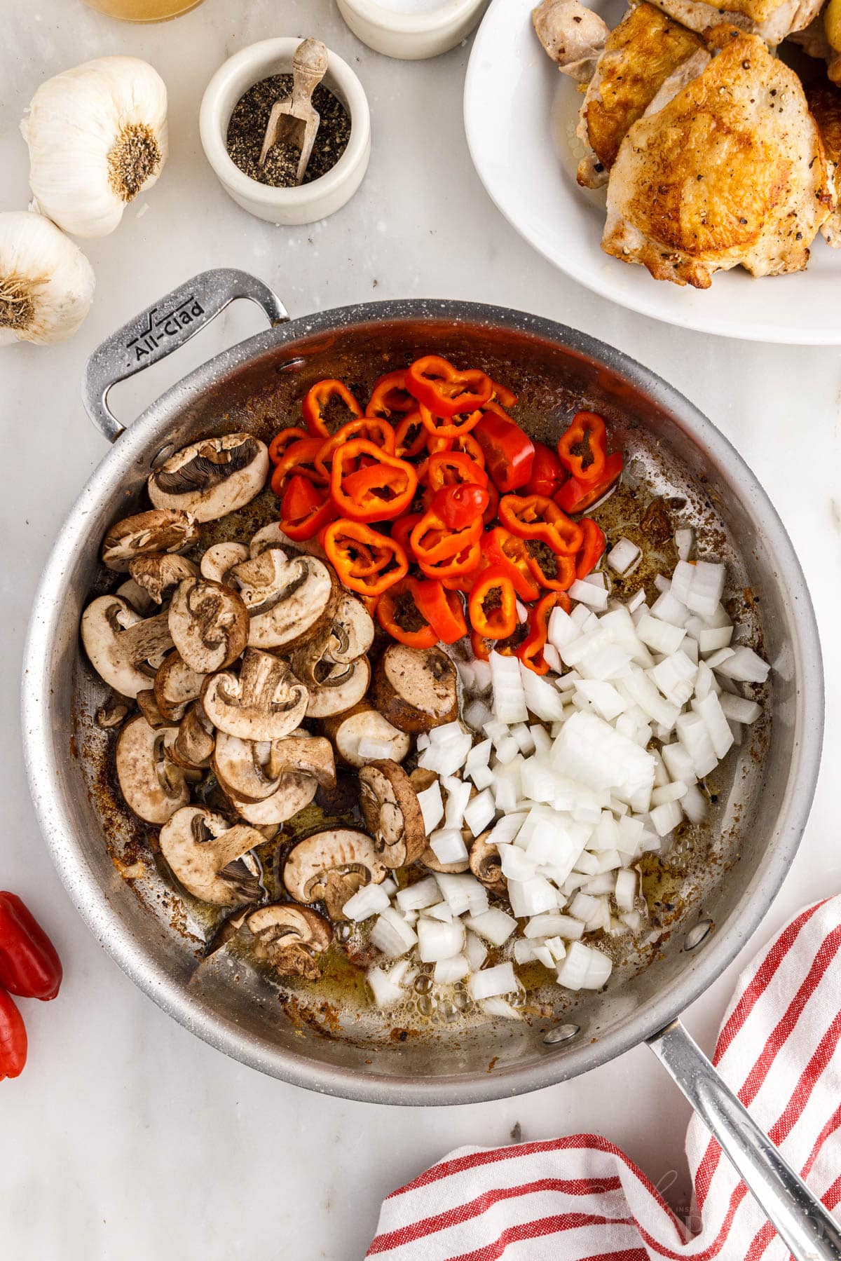 Chopped onions, red bell peppers, and mushrooms added to the cooking skillet with chicken set on a plate to the side.