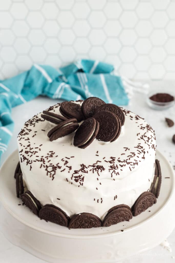 Oreos on top of ice cream cake and half oreos around bottom border with white frosting and chocolate sprinkles on white plate with aqua linen