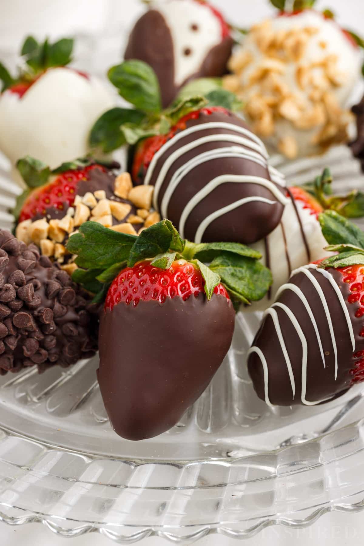 A glass platter of chocolate covered strawberries, some with dark or white chocolate, some also coated in crushed peanuts or mini chocolate chips.