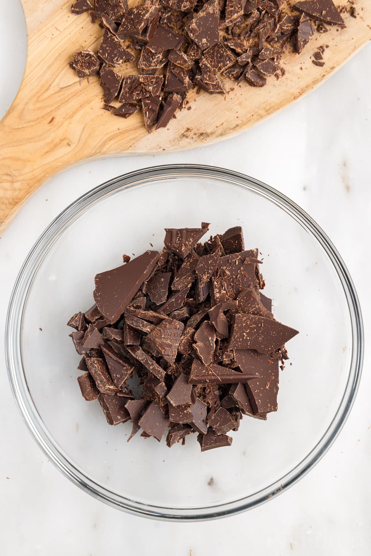 Chopped chocolate in glass bowl next to cutting board with chocolate pieces.