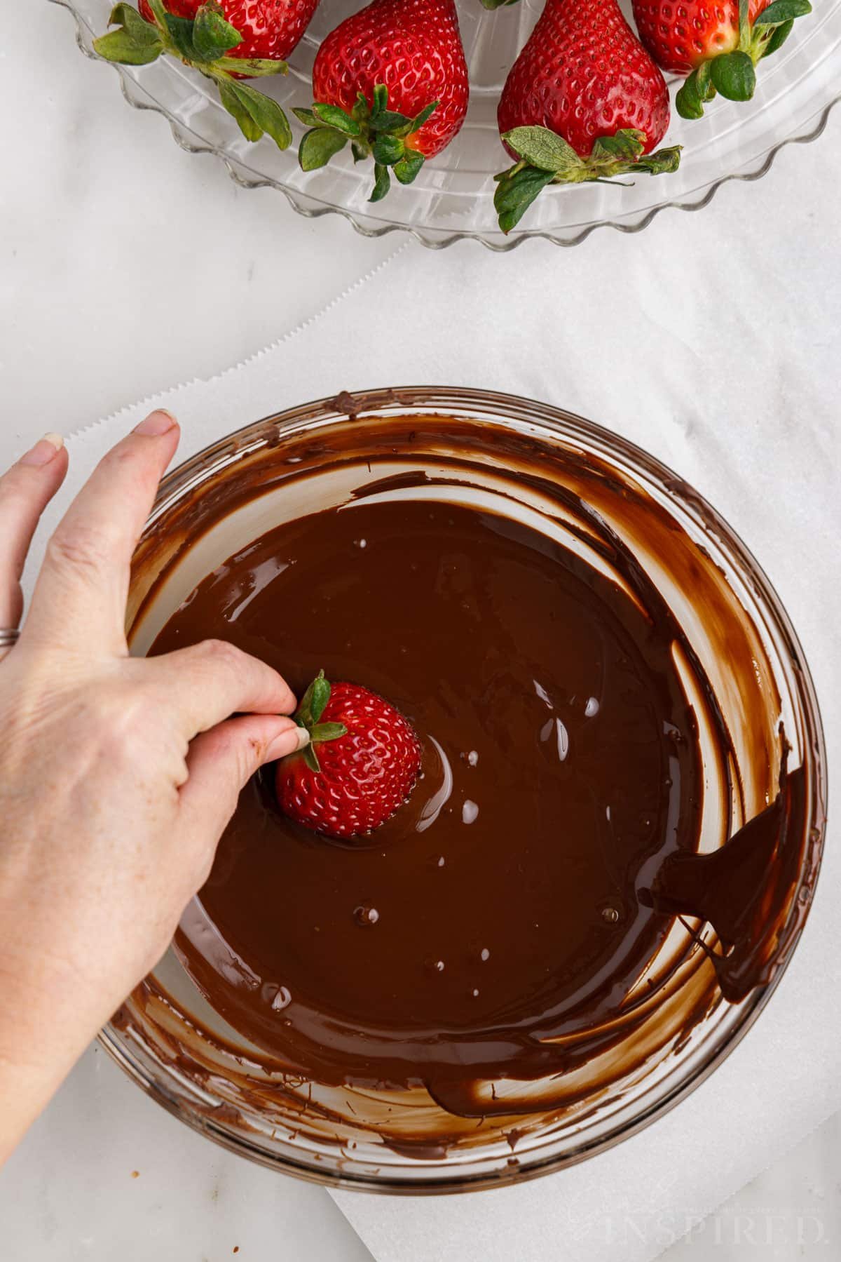 Holding a strawberry by its stem and dipping it into the bowl of melted chocolate.