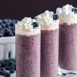 three tall glasses filled with blueberry kale smoothie and blueberries in white basket and bananas in the background