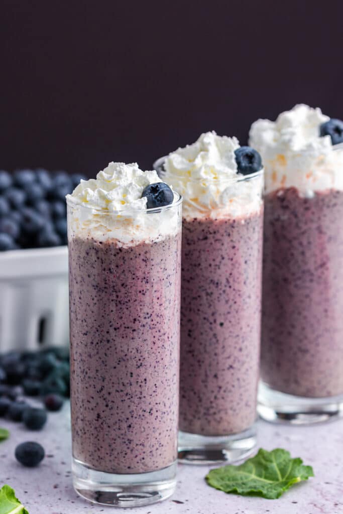 three tall glasses filled with blueberry kale smoothie and blueberries in white basket and bananas in the background