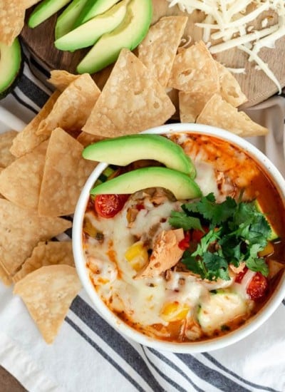 white soup bowl with cheese, avocado, and cilantro-topped soup, tortilla chips, and grated cheese on a striped dish towel