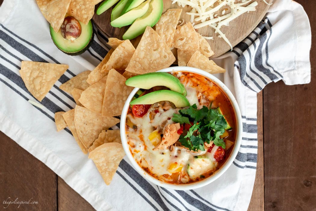 white soup bowl with cheese, avocado, and cilantro-topped soup, tortilla chips, and grated cheese on a striped dish towel