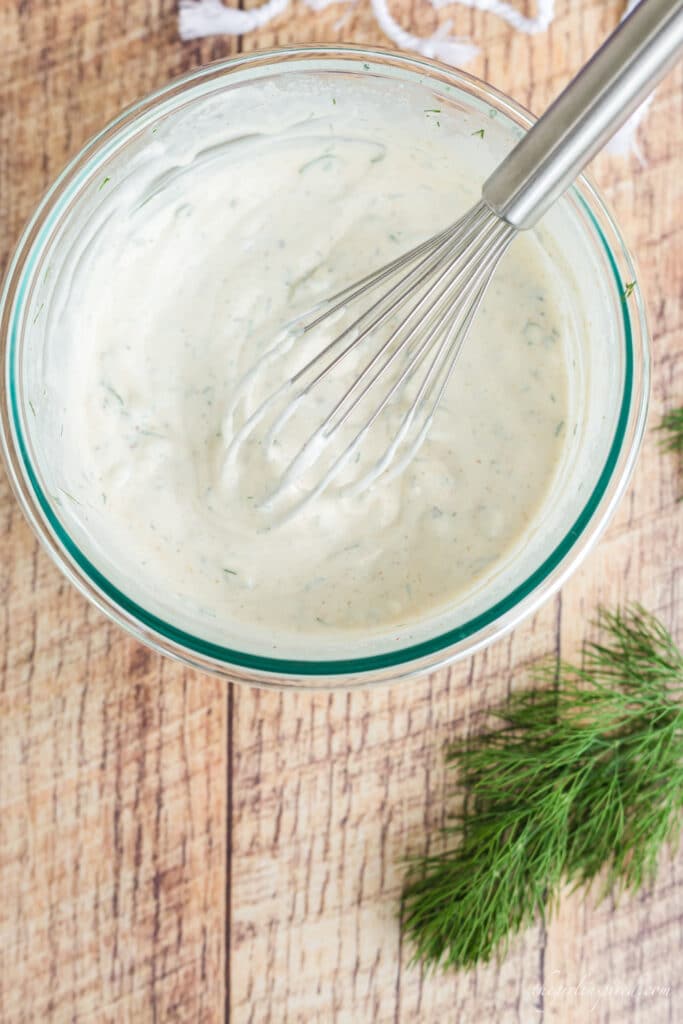 glass bowl of dill dip with wire whisk and sprig of fresh dill