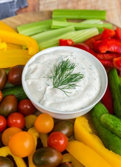 white bowl of dill dip with tomatoes, colored bell peppers, and celery on wooden cutting board