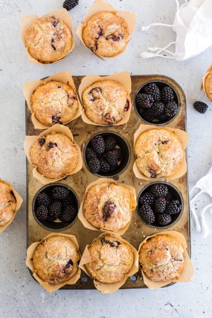 overhead view of blackberry muffins in muffin tin with some of the muffin spots filled with blackberries and a white linen