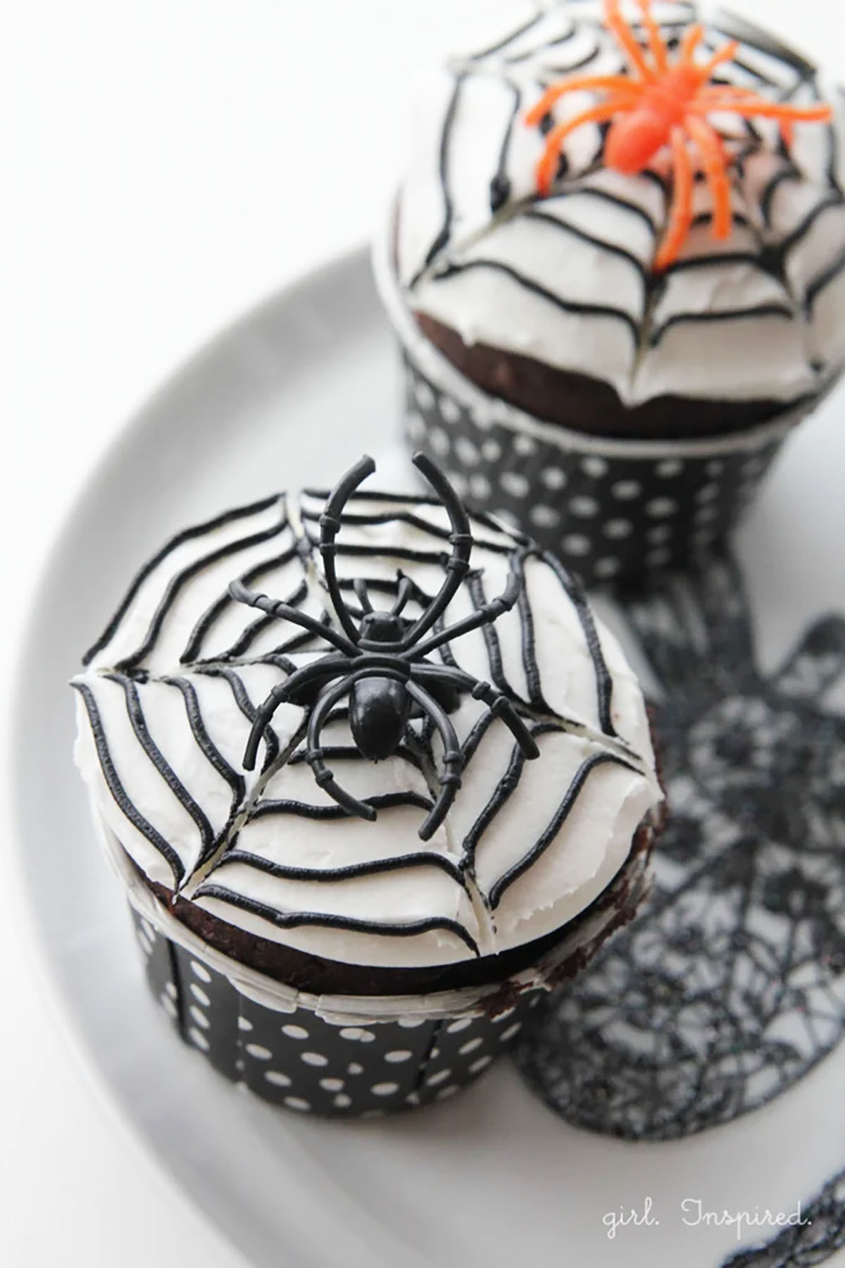 Two cupcakes with white icing and black spiderweb design, with plastic spiders on top.