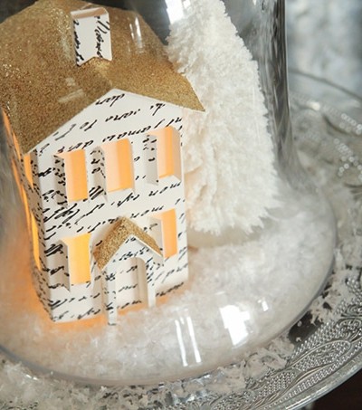 miniature paper house with gold roof, snow and white tree on glass plate with glass cloche over the top