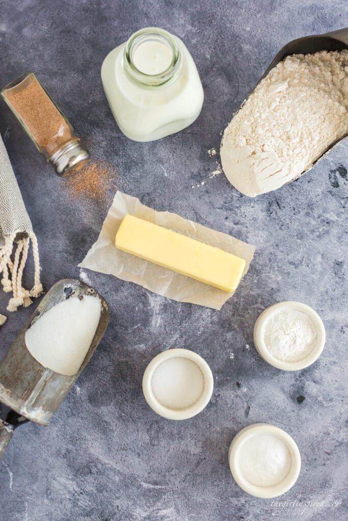 Buttermilk Scones ingredients: jar of buttermilk, cinnamon and sugar in a shaker, flour and sugar in metal scoops, butter, baking powder, soda, and salt in small bowls