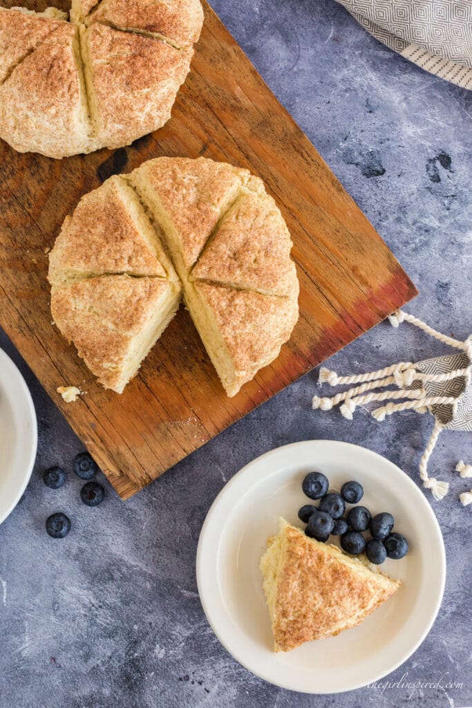 Scone rounds on wooden board with one scone triangle removed onto plate with blueberries