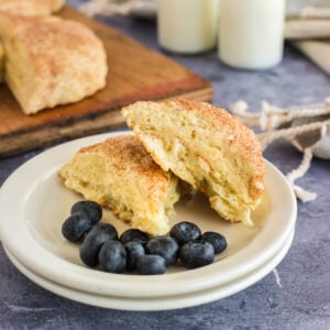 two buttermilk scones stacked on white plates with blueberries, milk and wooden board with scones in background