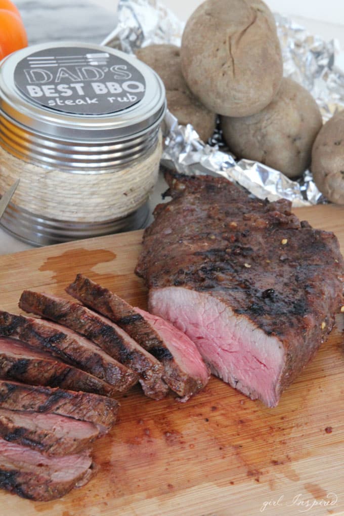 sliced steak on wooden cutting board with spice rub container and baked potatoes in background