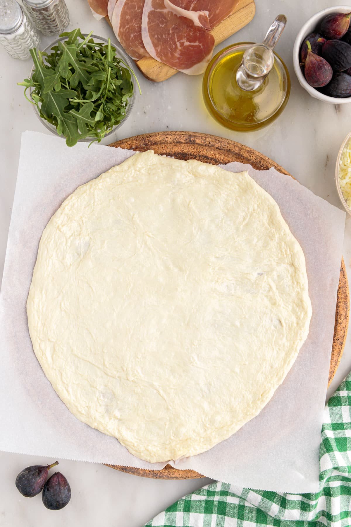 Rolled out pizza dough on parchment paper, with bowl of arugula, figs, olive oil, and green towel.