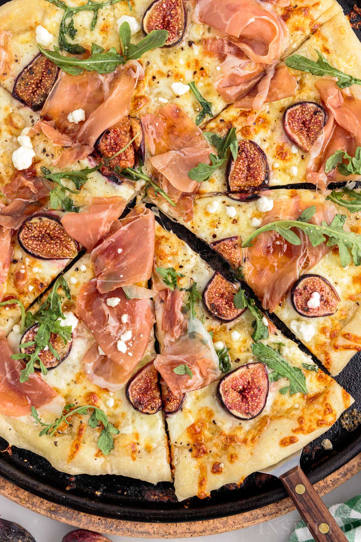 Overhead of fig prosciutto pizza on cast iron pan, cut into pie slices with serving spatula.