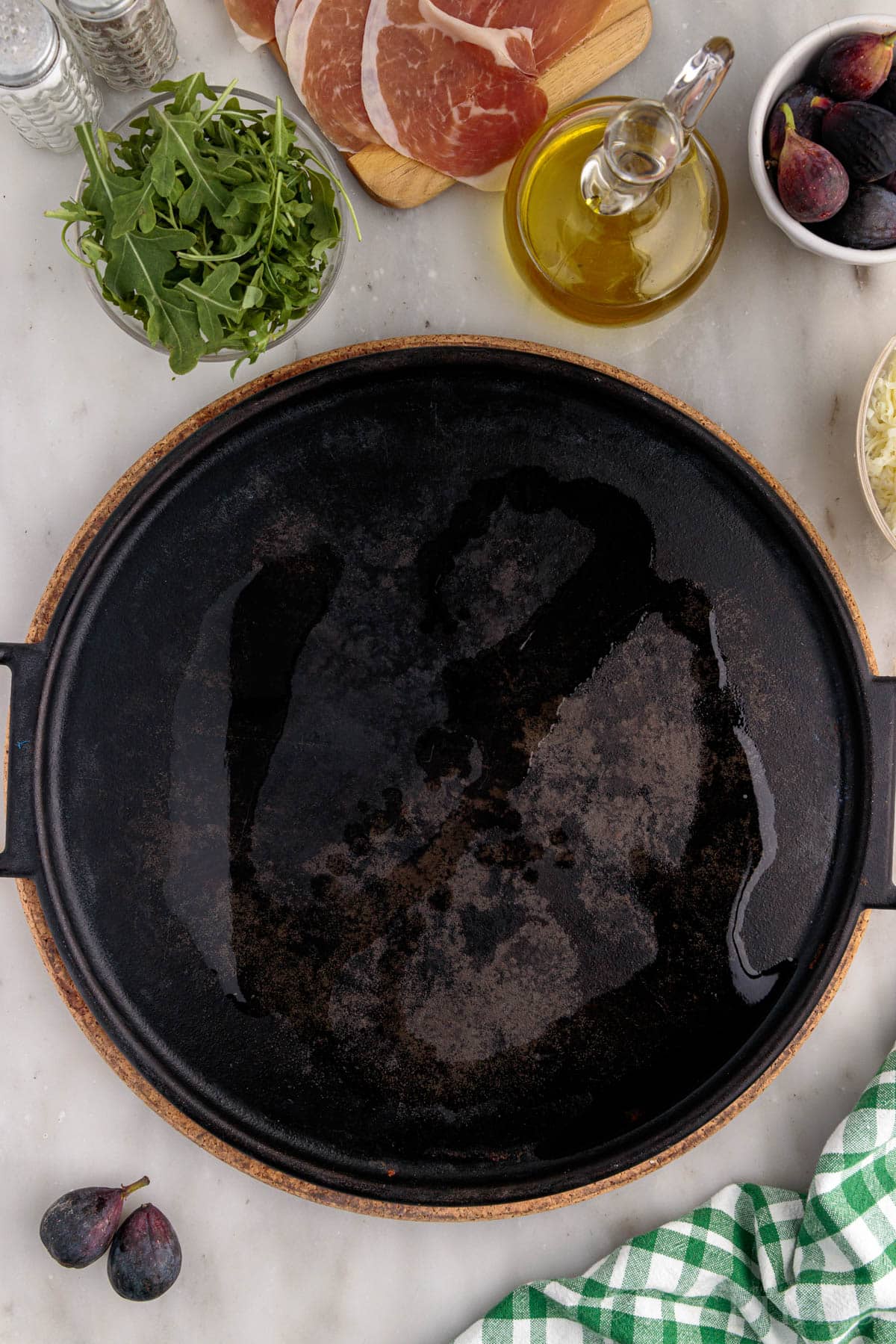 Olive oil on cast iron pizza pan, with bowl of aruglua, bowl of figs, olive oil, green towel.