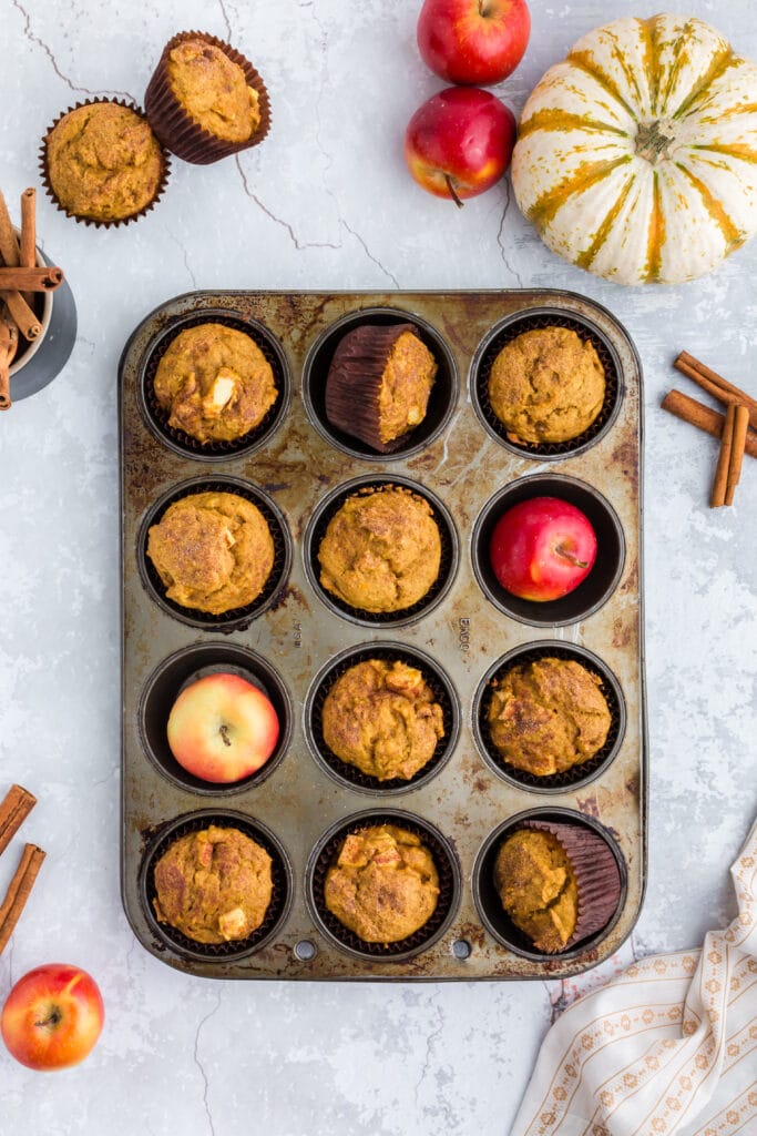 overhead of pumpkin apple muffins in muffin tin with red apples, pumpkins, and cinnamon sticks around