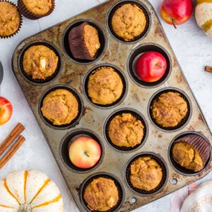 overhead of pumpkin apple muffins in muffin tin with red apples, pumpkins, and cinnamon sticks around