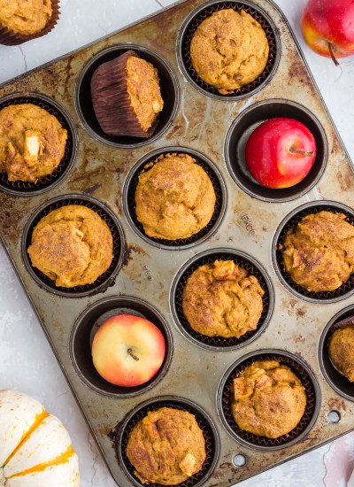 overhead of pumpkin apple muffins in muffin tin with red apples, pumpkins, and cinnamon sticks around