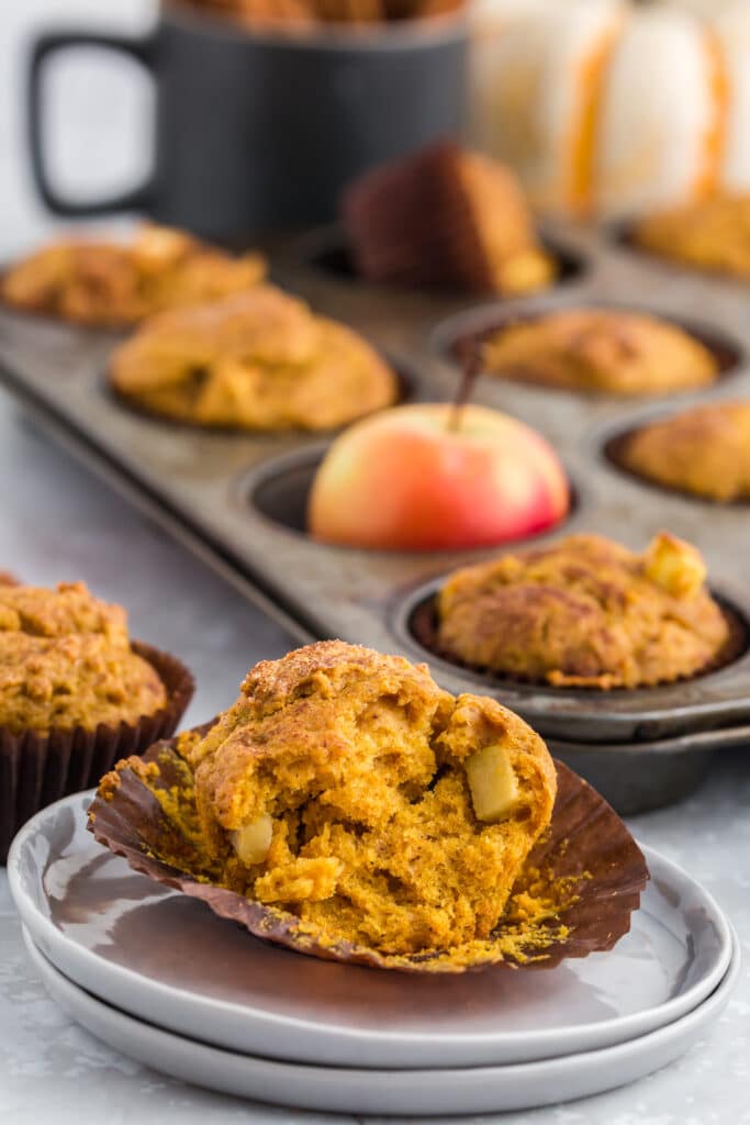 pumpkin apple muffin split open on plate in front of muffin tin with red apples, pumpkins, and cinnamon sticks