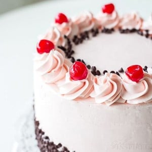 side view of pink frosted cake decorated with cherry buttercream swirls, maraschino cherries, and mini chocolate chips.