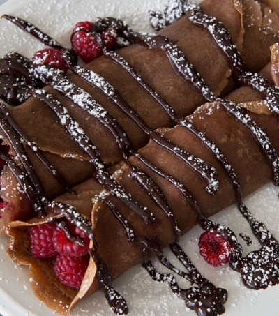 Chocolate crepes with raspberries inside, drizzled with chocolate sauce, sprinkled with powdered sugar, served on a white plate.