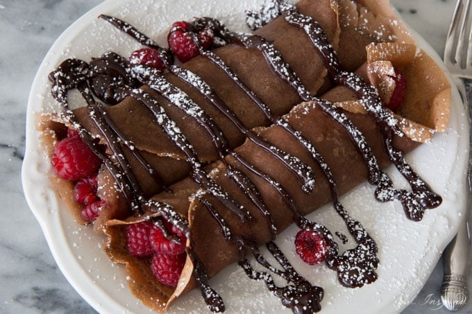 Chocolate crepes with raspberries inside, drizzled with chocolate sauce, sprinkled with powdered sugar, served on a white plate.