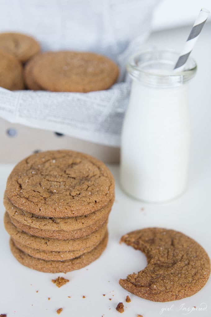 Ginger cookies stacked in newsprint-lined tin, cookies and jar of milk on white table in front of tin